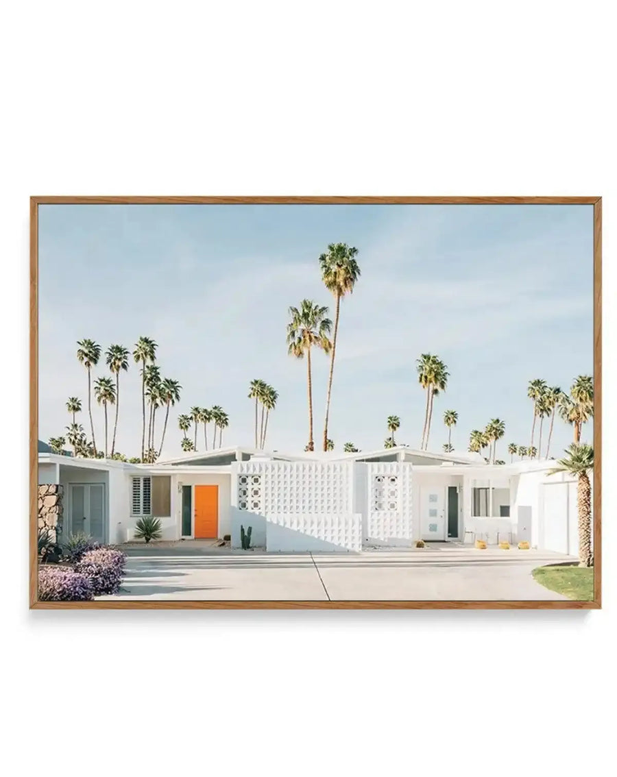 Framed canvas art print depicting a mid-century modern white single-story house with an orange front door, white geometric lattice wall panels, and minimal landscaping including purple flowering shrubs. Tall palm trees with green fronds line the clear blue sky behind the flat-roofed residence. The photograph-style illustration captures a desert modernist aesthetic in soft, naturalistic colors. Natural wood frame with white border around the canvas edges.