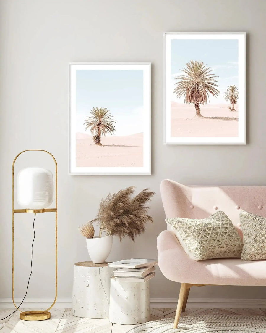 Lifestyle shot of a minimalist living room featuring two framed art prints displayed on a light grey wall above a blush pink sofa. The art prints are desert landscape photographs, each depicting a solitary palm tree with light brown fronds against a pale blue sky and soft pink sand. Both prints have white frames and white borders. The room includes a gold floor lamp with a white cylindrical shade, a white ceramic vase with dried pampas grass, and a stack of books on a white cylindrical side table.