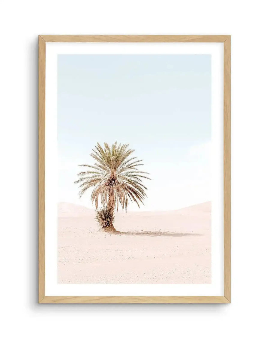 Framed photography art print of a solitary palm tree in a desert landscape, with a natural oak frame and white border. The composition features a single palm tree with light green and brown fronds, standing on soft, pale pink sand dunes under a clear, light blue sky.