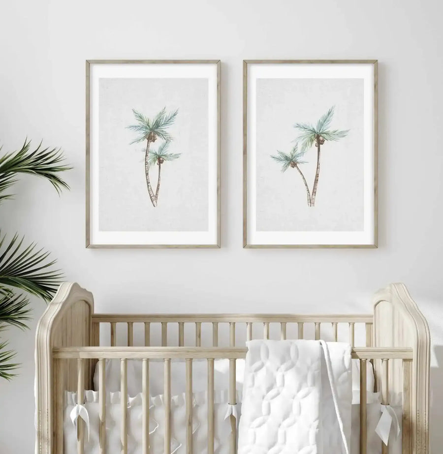 Nursery:
Two framed art prints featuring watercolor palm trees displayed above a light wood crib in a neutral-toned nursery. Each print depicts two palm trees with light blue-green fronds and brown trunks against a soft white background, with natural wood frames and white borders. The crib is adorned with white bedding and a quilted white blanket.