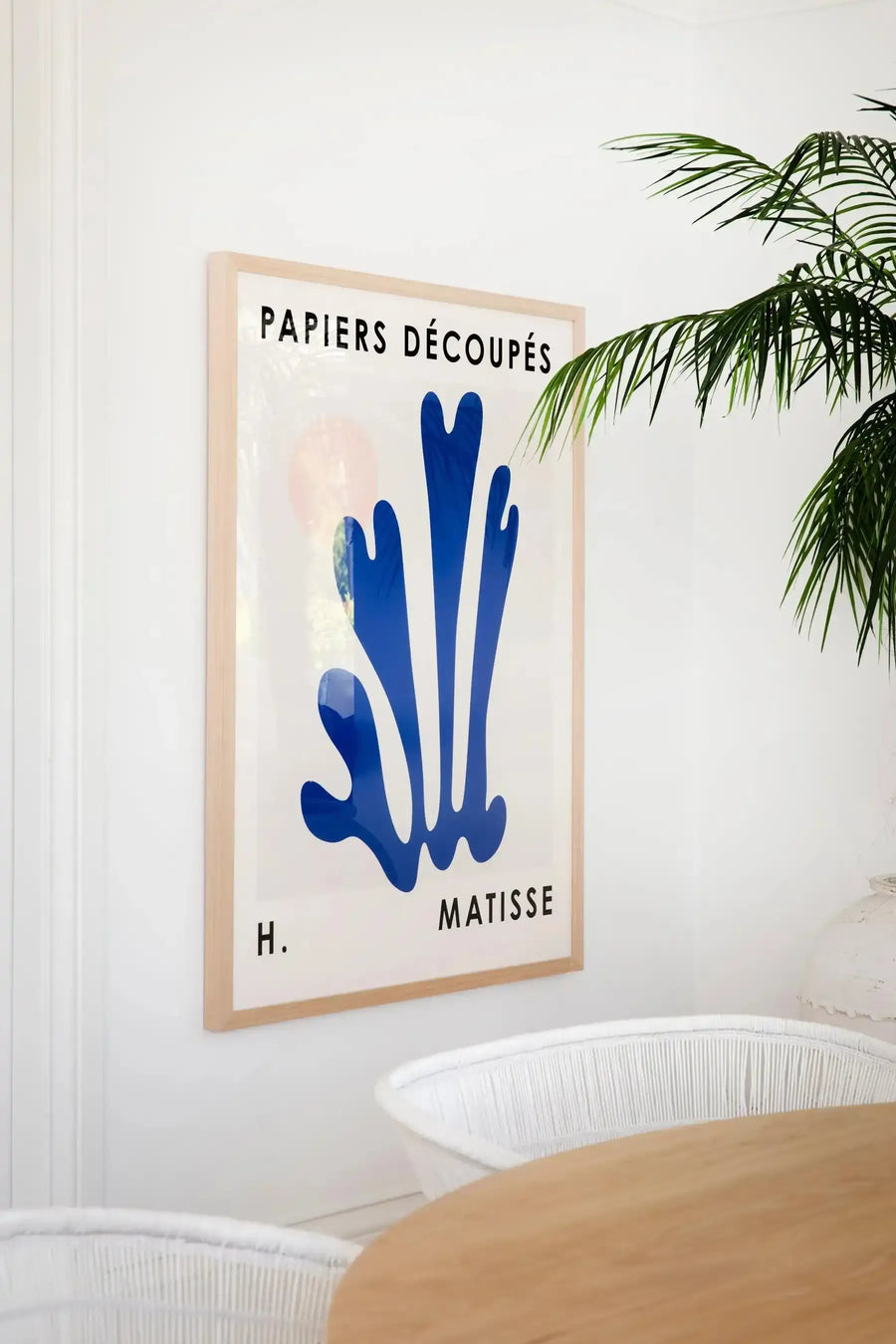 Lifestyle shot of a framed art print displayed on a white wall in a bright, minimalist dining room. The print features a large, abstract blue shape resembling seaweed or coral, with the text "PAPIERS DÉCOUPÉS" at the top and "H. MATISSE" at the bottom, all in black. The print is in a natural oak frame with a white border. A large green palm plant is visible in the top right corner, and a round wooden table with two white woven chairs is in the foreground.