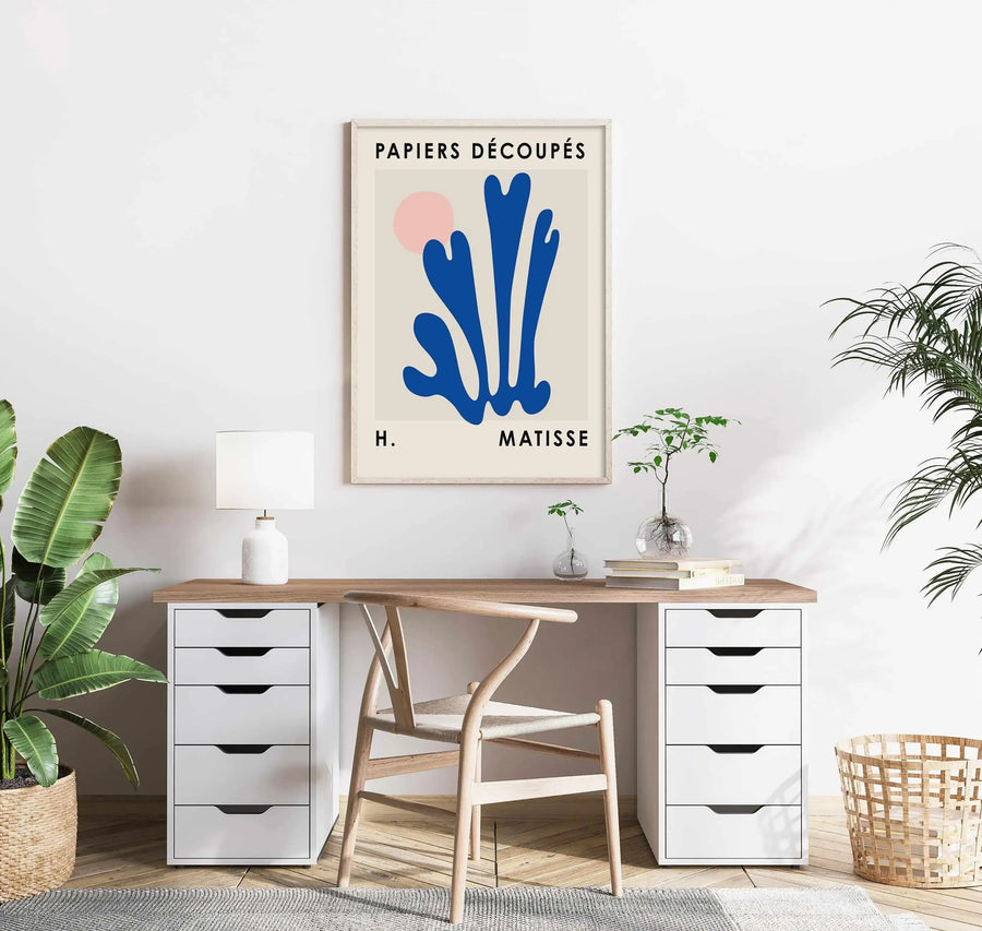 Home Office:
"Large framed art print displayed above a wooden desk with white drawers in a bright home office. The print features a large abstract blue shape with a small pink circle on a cream background, with a natural wood frame and white border. The room includes a large potted plant in a woven basket, a white lamp, and a wooden chair."