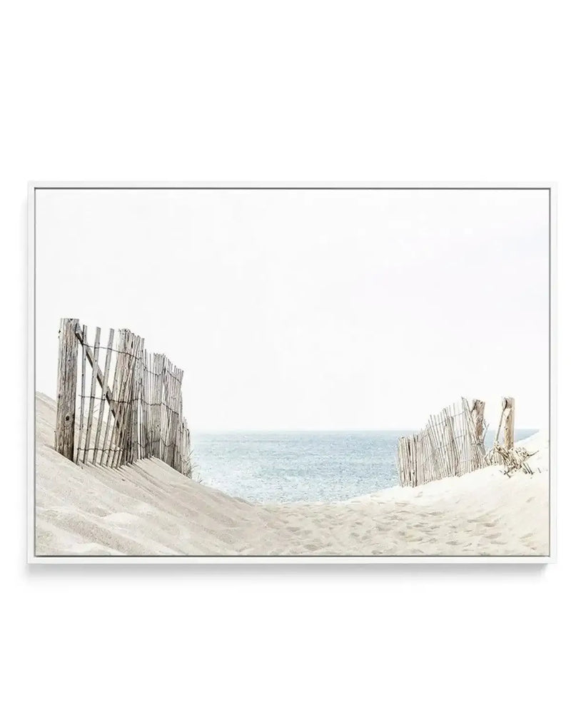 Framed canvas art print featuring a serene beach scene with weathered wooden dune fences on pale sand in the foreground, calm turquoise ocean and misty horizon in the background. The photograph has soft, muted tones with beige, gray, and pale blue hues. The canvas is mounted in a slim gray frame with a white border.