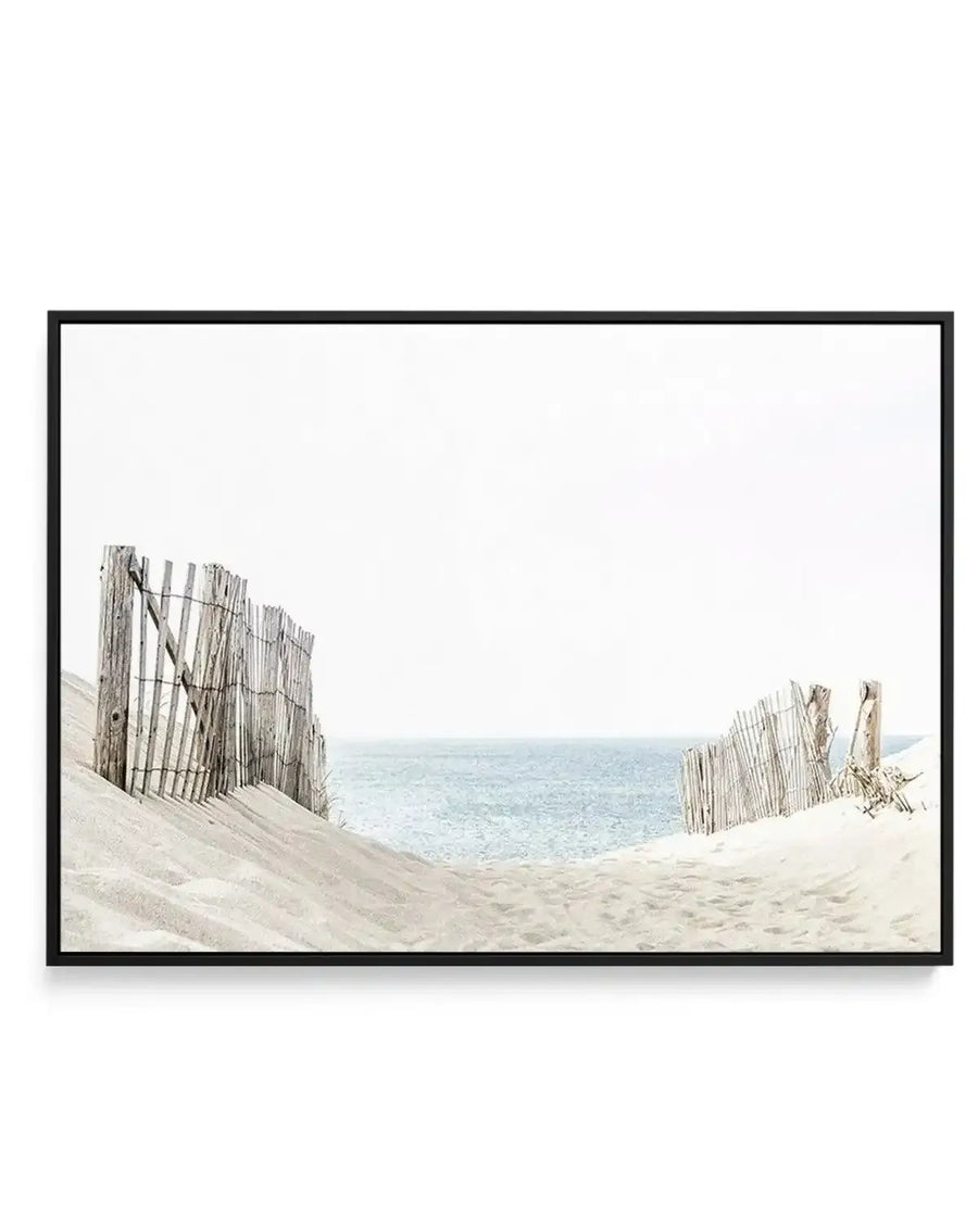 Framed canvas art print featuring a coastal landscape photograph. Weathered wooden sand fence sections frame a view of pale sandy beach sloping toward calm turquoise ocean under a hazy sky. The composition uses the fence posts as natural framing elements on left and right sides. Soft, muted tones of beige, gray, and pale blue create a serene, coastal mood. The photograph has a clean, naturalistic quality. Black frame with white border surrounds the canvas.