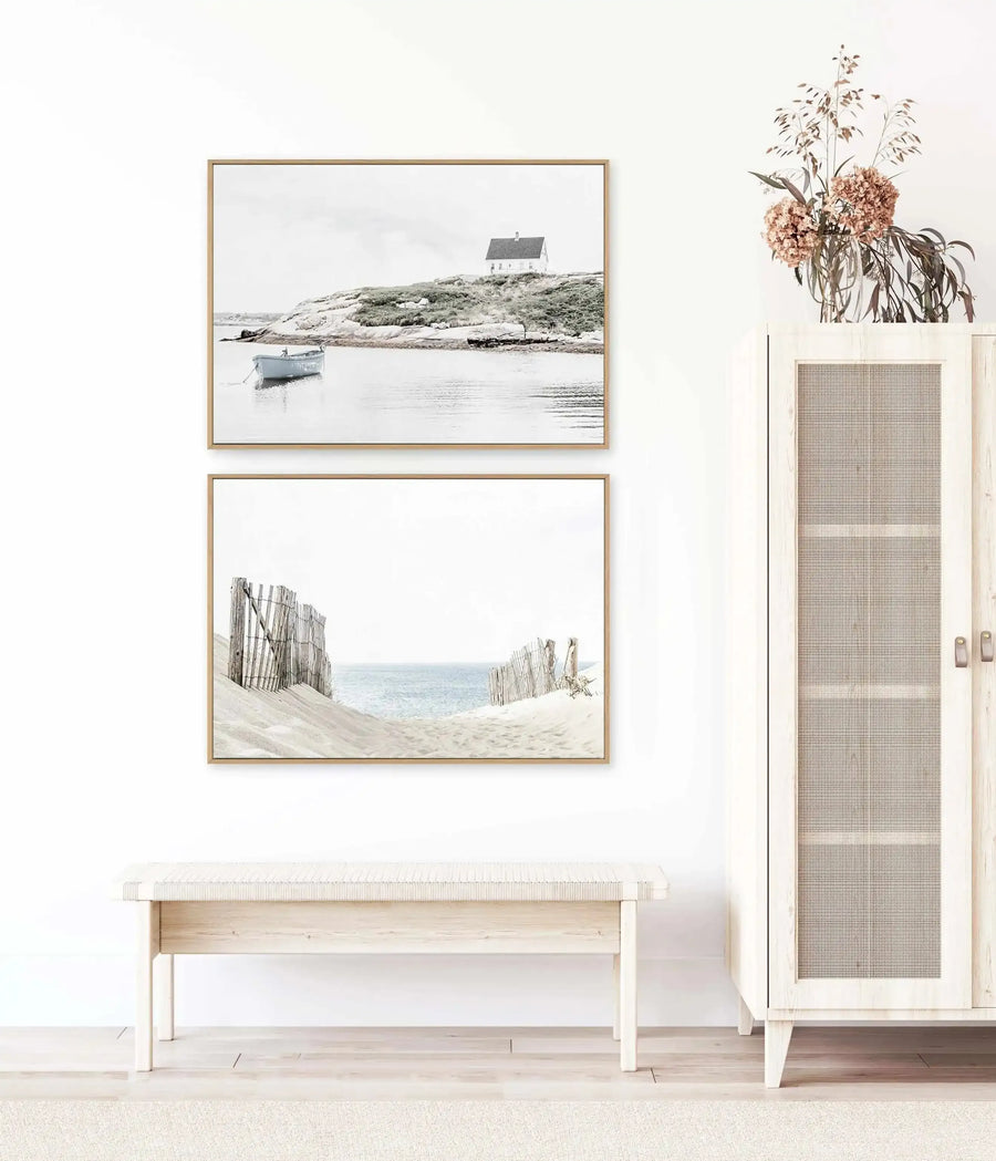 Lifestyle shot of two framed canvas art prints displayed above a light wood bench in a minimalist room with a light wood cabinet. The top print is a black and white photograph of a small white house on a grassy hill overlooking a calm body of water with a small boat. The bottom print is a black and white photograph of a sandy beach path with a weathered wooden fence leading to the ocean. Both prints have natural wood frames and white borders. A vase of dried flowers sits on the cabinet.