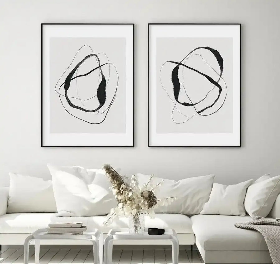 Lifestyle shot of two framed art prints displayed above a white sectional sofa in a minimalist living room. Each print features abstract, organic shapes in thick black lines and solid black fills on a white background, with black frames and white borders. The room includes a white coffee table with books, a clear glass vase with dried pampas grass, and a grey throw blanket.