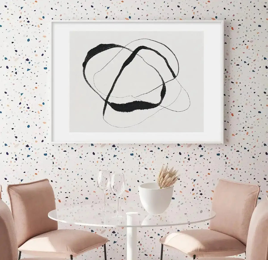 Dining Room:
Large framed art print displayed above a round glass dining table with two blush pink upholstered chairs in a modern dining room. The art print features bold, abstract black brushstrokes forming an intertwined, organic shape on a white background, with a white frame and white border. The wall is covered in white terrazzo-patterned wallpaper with multi-coloured flecks.