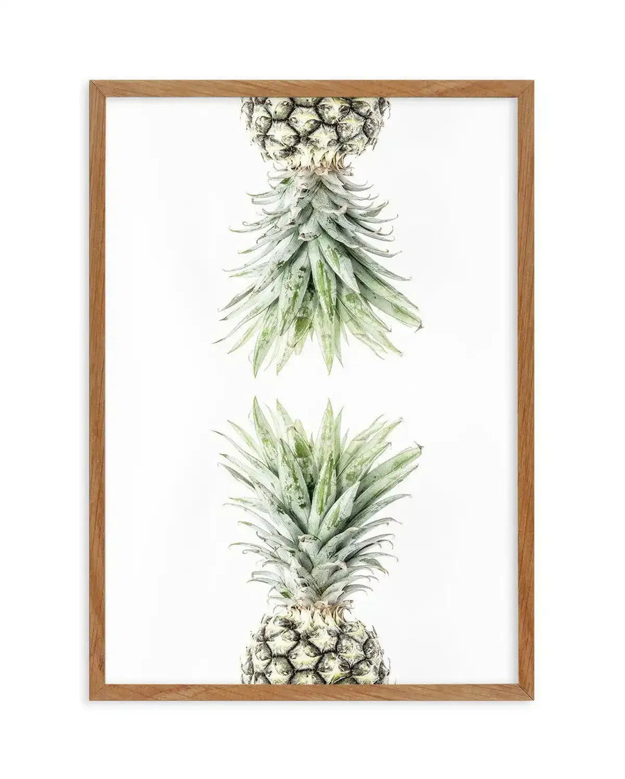 Framed art print displaying two pineapples in vertical composition against a white background. The top pineapple is shown inverted with its textured fruit at top and long, pointed green leaves cascading downward. The bottom pineapple is oriented normally with layered green leaves at top and bumpy fruit base. Rendered in detailed watercolor illustration with soft greens, pale yellows, and warm browns creating a naturalistic botanical style. Natural wood frame with light tan finish completes the presentation.