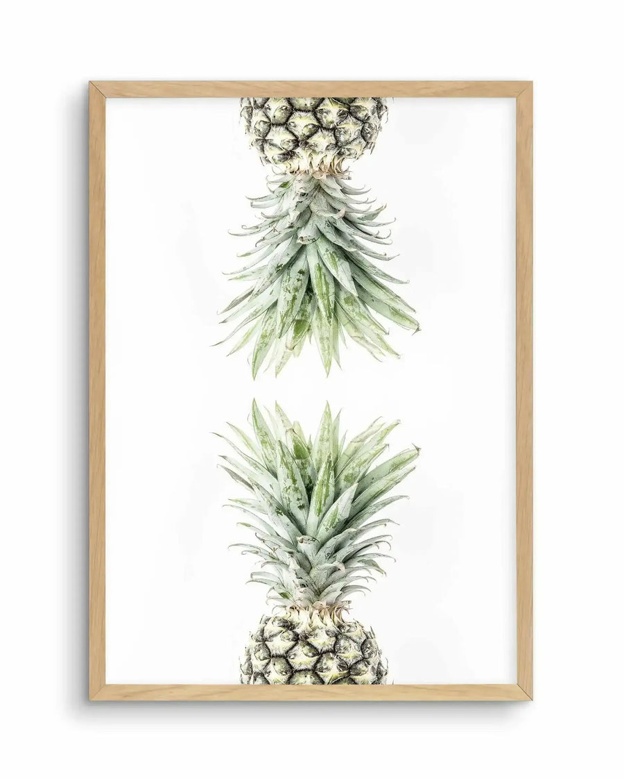 Framed art print featuring watercolor botanical illustration of two pineapples arranged vertically on white background. Top pineapple shown upside down displaying detailed crown of long, pointed green leaves with subtle gray-green shading. Bottom pineapple shown right-side up with textured fruit body in cream and tan tones. Natural wood frame with white border surrounding the artwork.