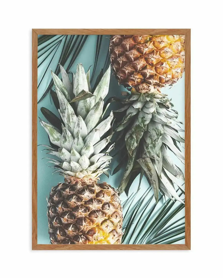 Framed photography art print featuring two pineapples and palm fronds on a light blue background, displayed in a natural wood frame with white border. The composition shows one pineapple upright in the foreground and another inverted in the background, with the green palm leaves peeking out from behind.