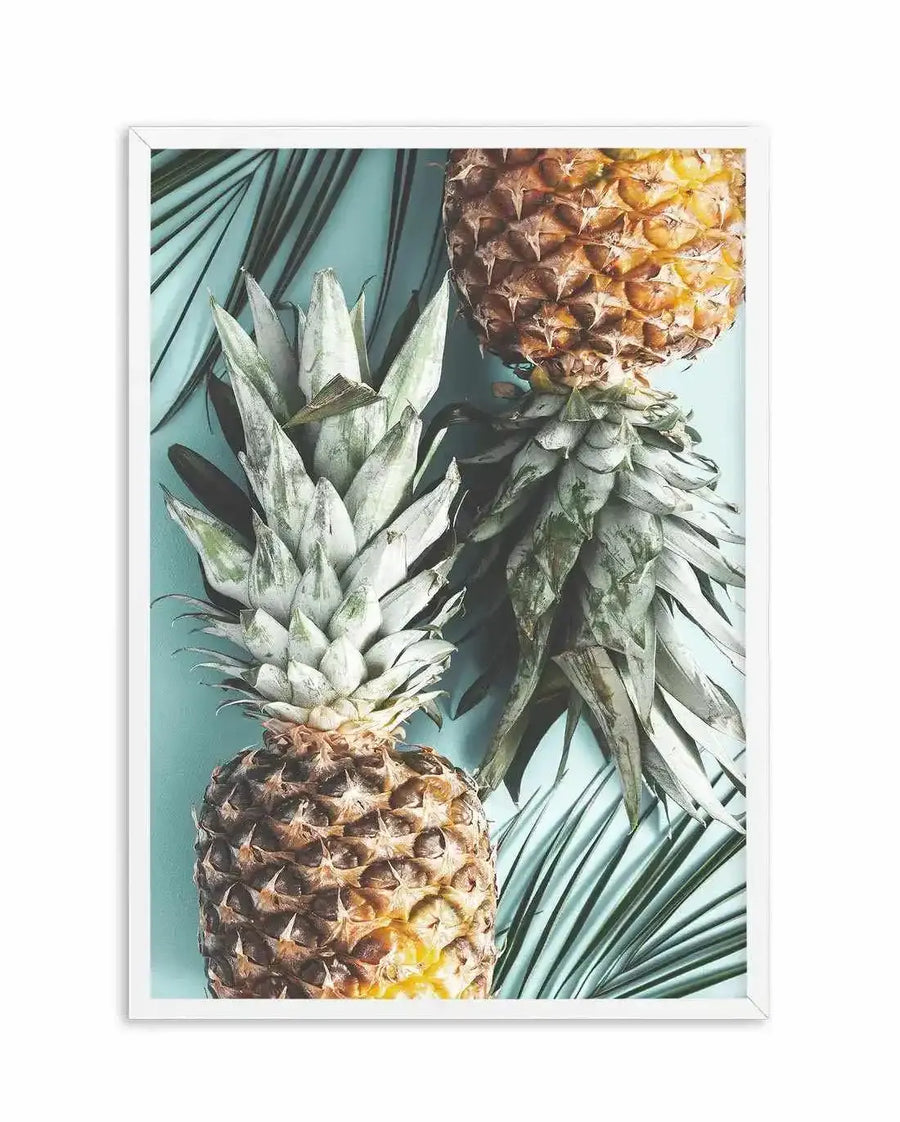 Framed photography art print featuring two pineapples and palm fronds on a light blue background, with a white frame and white border. The composition shows one pineapple upright in the lower left, with its spiky green crown extending upwards. The second pineapple is positioned above and to the right, inverted, with its crown extending downwards. Dark green palm fronds are visible in the background, creating a tropical and fresh mood.