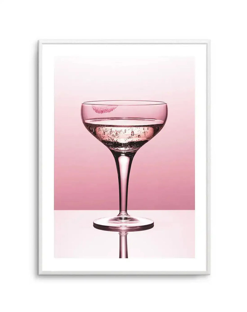 Framed photography art print of a coupe glass filled with sparkling pink liquid and a bright pink lipstick kiss mark on the rim, set against a soft blush pink background with a white surface reflecting the glass, displayed in a white frame with white border.
