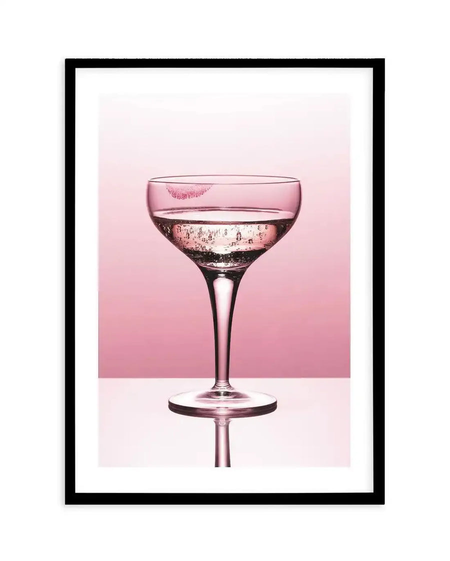 Framed photograph of a coupe glass with a pink lipstick kiss mark on the rim, filled with sparkling pink liquid and bubbles, set against a soft pink background with a reflective white surface below, displayed in a black frame with a white border.