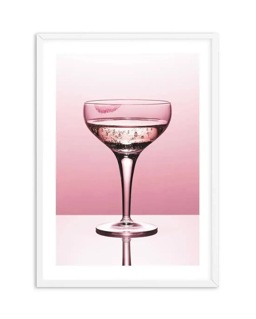 Framed photography art print featuring a coupe glass filled with sparkling pink liquid and a bright pink lipstick kiss mark on the rim, set against a soft blush pink background with a reflective white surface below, displayed in a white frame with a white border.