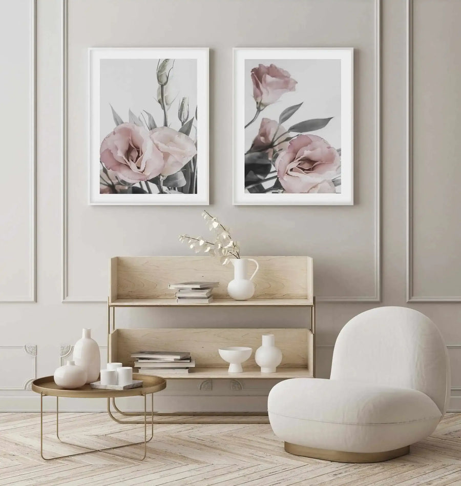 Framed art print displaying two vertical compositions of dusty rose carnations with delicate green foliage against a soft gray background. Left frame shows clustered blooms with unopened buds; right frame features individual flowers at varying heights. Both prints feature soft, watercolor-style rendering with muted tones. White frames with white borders hang above a light wood shelf unit in a minimalist room styled with cream furnishings, potted white ceramics, and a round brass side table.