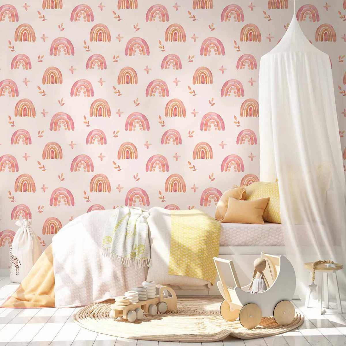 Nursery bedroom wall featuring Pink Pop Rainbows Wallpaper displayed behind a white canopy bed with yellow and peach bedding. The wallpaper pattern shows repeating hand-painted style rainbows in shades of dusty rose, coral, and terracotta with cream stripes, interspersed with small botanical leaf sprigs and plus sign motifs on a soft cream background. The illustration has a watercolor aesthetic with organic, gentle brushwork. Wooden toys and a white toy pram sit on a round rug in the foreground.