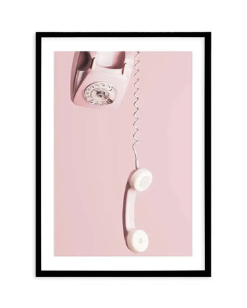 Framed photography art print of a vintage pink rotary telephone with the receiver hanging by its coiled cord against a solid light pink background, with black frame and white border. The telephone is positioned in the upper left corner, angled slightly downwards, revealing the circular dial with numbers and finger holes. The white receiver hangs vertically in the lower right, casting a soft shadow on the pink background.