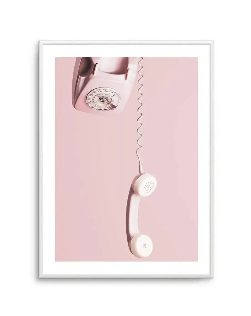 Framed photography art print featuring a vintage rotary telephone in a soft blush pink, with the receiver hanging down by its coiled cord against a solid light pink background. The telephone base is positioned in the upper left corner, showing the rotary dial with numbers and finger holes. The white receiver hangs vertically in the lower right, connected by a long, wavy white cord. The print is displayed in a white frame with a white border.