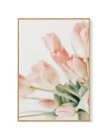 Soft-focus photograph of pale pink and white tulips with green stems arranged in a vertical composition. The delicate petals are rendered in blush and cream tones with coral-pink accents, creating a dreamy, impressionistic effect. The image is printed on canvas and framed in natural wood with a white border, presented against a neutral background.