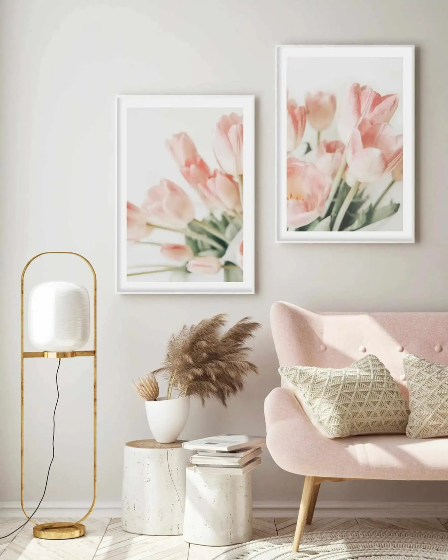 Lifestyle shot of two framed art prints displayed on a light grey wall above a blush pink sofa in a bright, minimalist living room. The prints feature soft focus photography of delicate pink tulips with green stems against a light background, with white frames and white borders. The room includes a gold floor lamp, a white ceramic vase with dried pampas grass, and two white side tables with books.