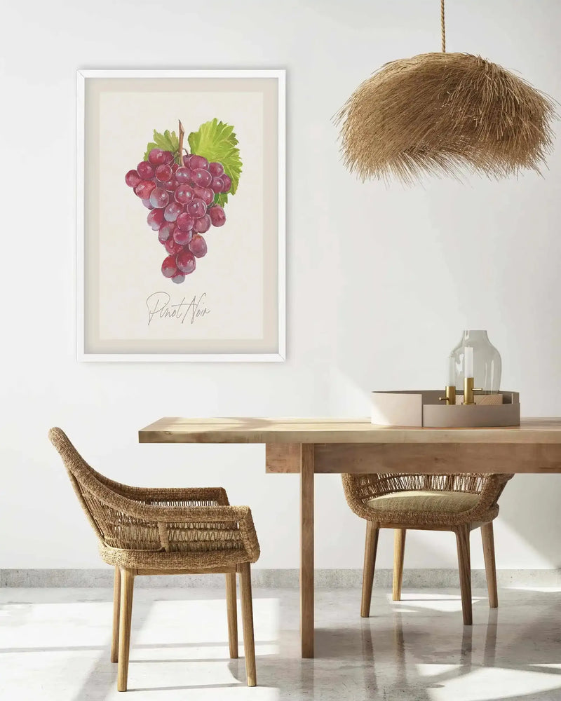 Lifestyle shot of a dining room featuring a framed art print of a bunch of red grapes with the text "Pinot Noir" in a script font, displayed on a white wall. The print has a white frame and a light cream border. Below the print is a light wood dining table with two woven wicker chairs. On the table, there is a decorative tray with a clear glass vase and two brass candle holders. A large, natural straw pendant light hangs from the ceiling above the table.