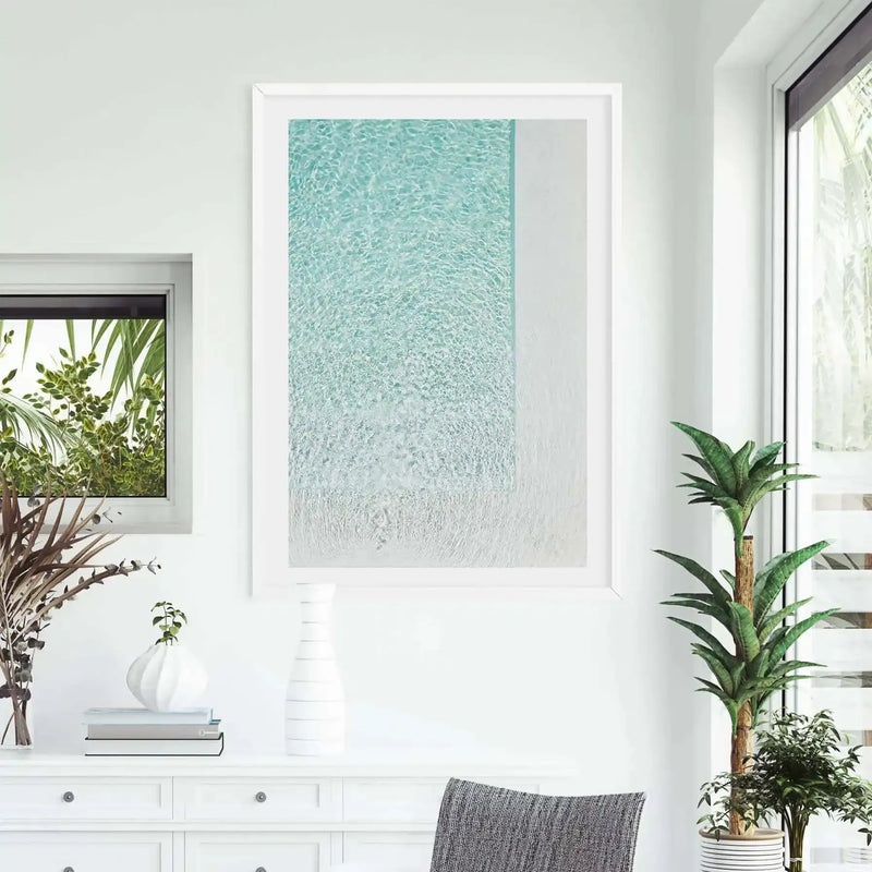 Lifestyle shot of a minimalist room featuring a large framed art print of a clear blue swimming pool with a white edge, viewed from above. The art print has a white frame and white border. The room has white walls and a white console table with decorative items, including a white vase with dried foliage, a stack of books, and a small plant. A tall green plant in a striped pot stands next to a large window on the right.