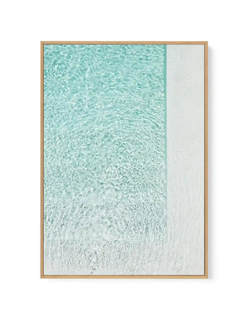Framed canvas art print featuring an aerial view of shallow turquoise water transitioning to white sandy beach. The composition shows rippling water patterns in varying shades of aqua and seafoam green in the upper two-thirds, gradually fading to pale cream and white sand at the bottom. Natural wood-toned frame with clean lines displaying the photograph-style artwork with white border.