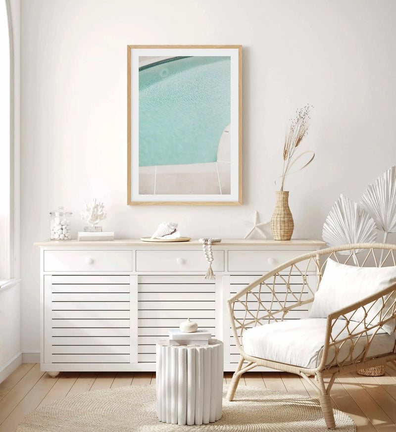 Lifestyle shot of a minimalist living room with a framed art print of a swimming pool. The print features a close-up, high-angle view of the turquoise water with gentle ripples and a light-colored pool edge, displayed in a natural wood frame with a white border. The room has white walls, light wood flooring, a white slatted cabinet with decor, and a rattan armchair with a white cushion.
