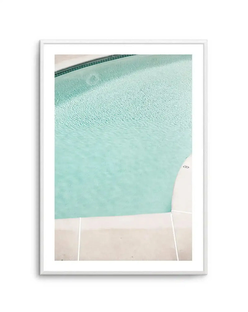 Framed photography art print of a swimming pool with clear, rippling turquoise water and a dark green tiled edge, with a white frame and white border. The composition shows the corner of the pool and surrounding light beige paving.