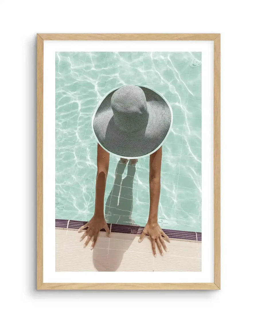 Framed photography art print of a person in a swimming pool, viewed from above, with a natural oak frame and white border. The person is wearing a wide-brimmed, light grey woven straw hat, with their arms resting on the pool edge. The water is a clear, light turquoise with rippling sunlight patterns on the surface and tiled bottom.