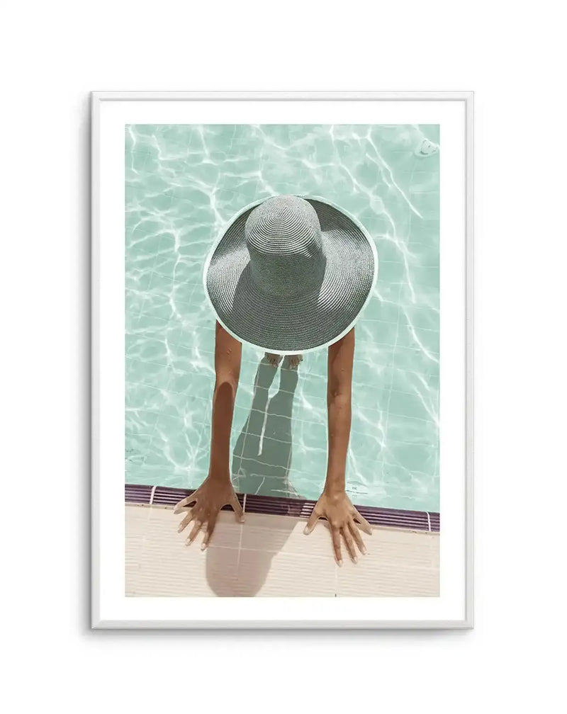 Framed photography art print of a person in a wide-brimmed straw hat sitting on the edge of a swimming pool. The person's back is to the viewer, with their arms resting on the pool edge and feet submerged in the clear, light blue water. The sun creates rippling reflections on the water and casts shadows of the person's arms and feet. The print is displayed in a white frame with a white border.