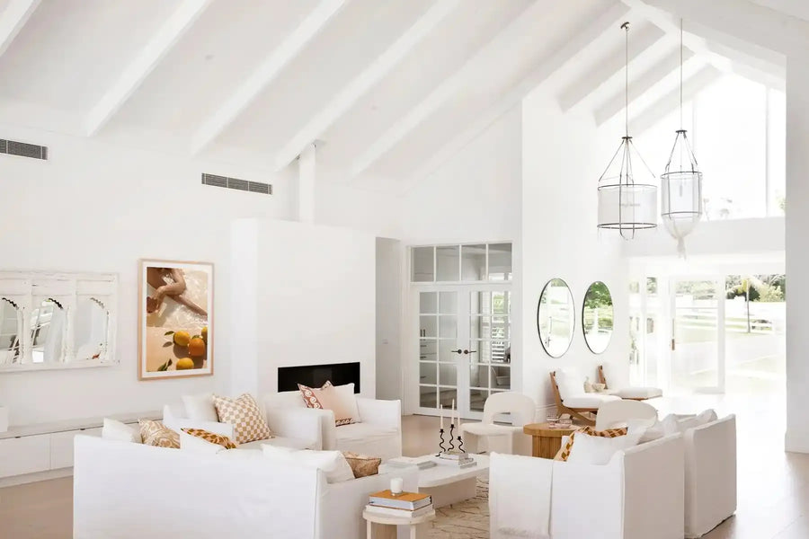 Lifestyle shot of a minimalist living room featuring the "Pool Time II" art print displayed above a white console table. The print is a framed photograph of a person's legs partially submerged in clear water, with several bright yellow and orange lemons floating on the surface. The art print is framed in natural wood with a white border. The living room has white walls, a vaulted ceiling with exposed white beams, and white slipcovered sofas with patterned throw pillows.