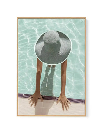 Framed canvas art print featuring an overhead view of a person leaning over a swimming pool edge, their face obscured by a large wide-brimmed hat with white and dark green stripes. The person wears a dark green and white checkered swimsuit. Clear turquoise pool water with rippling light patterns fills the background. The pool deck shows a dark striped border. Natural wood frame with white border surrounding the artwork.