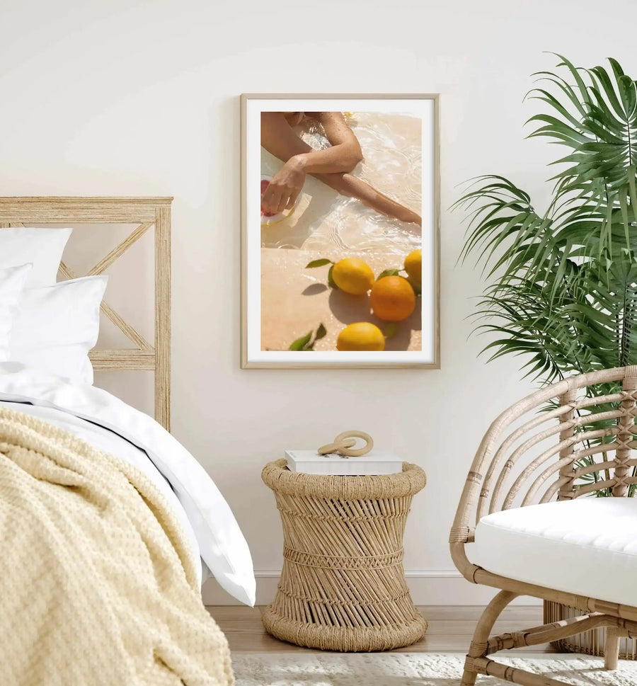 Lifestyle shot of a bedroom featuring a framed art print above a woven side table. The print, titled "Pool Time II," is a photograph of a person's legs and arm partially submerged in clear, shallow water on a light sandy beach, with several oranges and lemons scattered on the sand. The print has a natural wood frame and a white border. The bedroom includes a light wood headboard, white bedding with a cream knitted throw, a large green palm plant, and a rattan armchair with a white cushion.