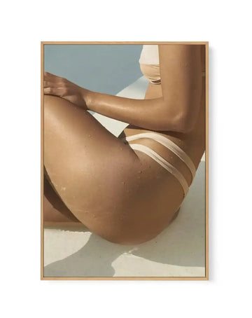 Framed canvas print featuring a close-up, cropped photograph of a person in a light beige bikini sitting by a pool. The person's skin is tanned and covered in water droplets, with strong sunlight creating shadows on the white pool edge. The background is a soft blue, suggesting water or sky, and the image is framed in natural oak.