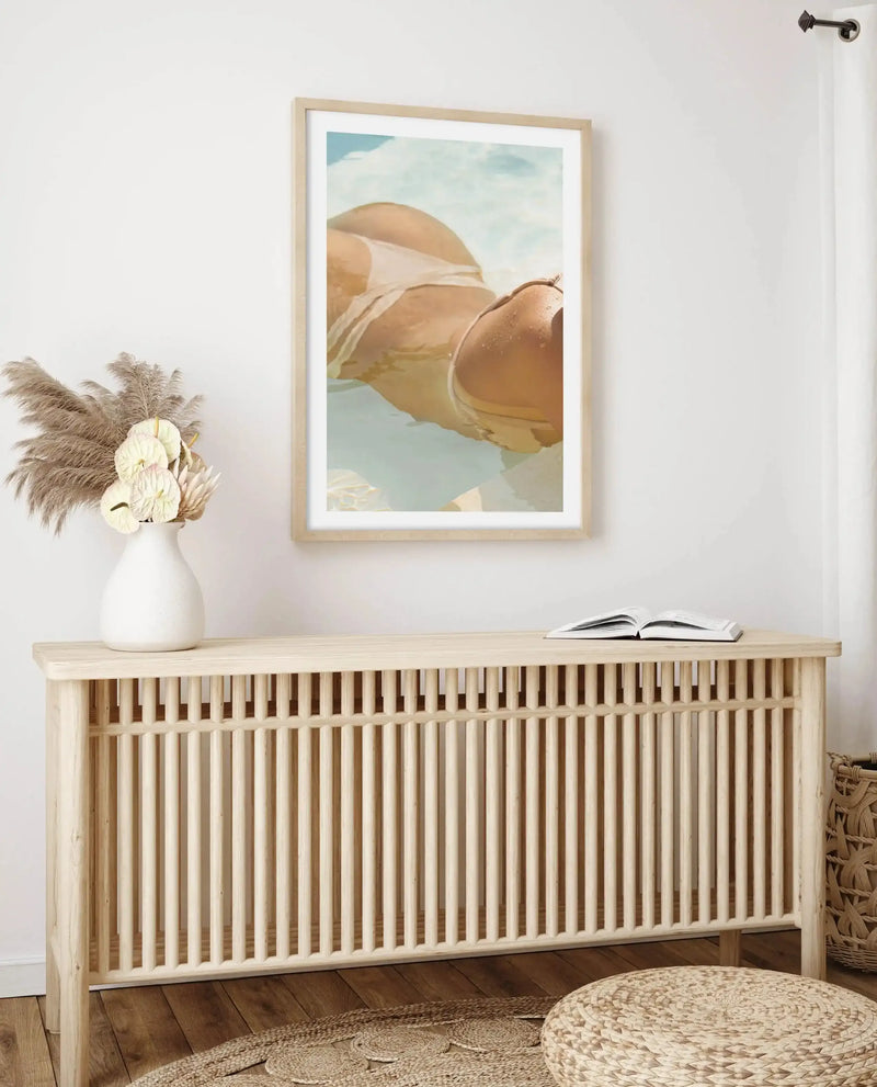Lifestyle shot of a framed art print displayed above a light wood console table in a minimalist room. The print features a close-up, overhead photograph of a person in a white bikini floating in clear blue water, with their back and legs visible. The print is in a natural wood frame with a white border. On the console table, a white vase holds dried pampas grass and white anthurium flowers, and an open book rests on the right side. A woven jute rug and a round woven pouf are on the wooden floor.