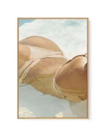 Canvas art print titled "Pool Time IV" featuring an illustration of a woman's torso and legs in a tan-colored bikini, partially submerged in clear turquoise water. The figure is rendered in soft, painterly strokes with warm peachy and golden tones. White fabric drapes across the upper portion. The background shows a pale blue sky with white clouds and sandy terrain. The composition has a dreamy, relaxed quality with soft lighting. The canvas is mounted in a natural wood frame with a white border.