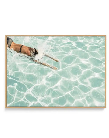 Framed canvas art print featuring an overhead photograph of a person swimming freestyle stroke in clear turquoise water with white ripple patterns. The swimmer wears a tan-colored swim cap and brown arm band, captured mid-stroke with one arm extended forward. The water's surface shows intricate light refraction patterns in white against soft aqua and seafoam tones. Natural wood-tone frame with horizontal orientation.