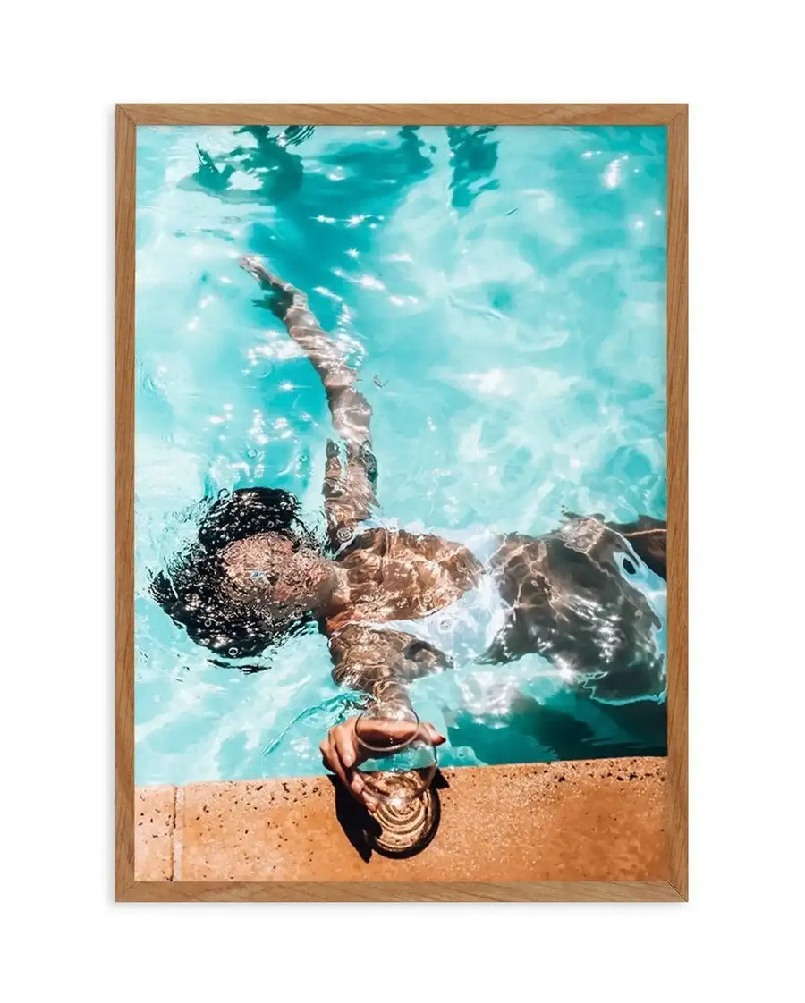 Framed photography art print of a person floating in a swimming pool, viewed from above, with a natural wood frame and white border. The person is submerged in bright turquoise water, holding a drink in a clear glass with a gold base, resting on the pool's edge. Sunlight creates sparkling reflections on the water's surface.
