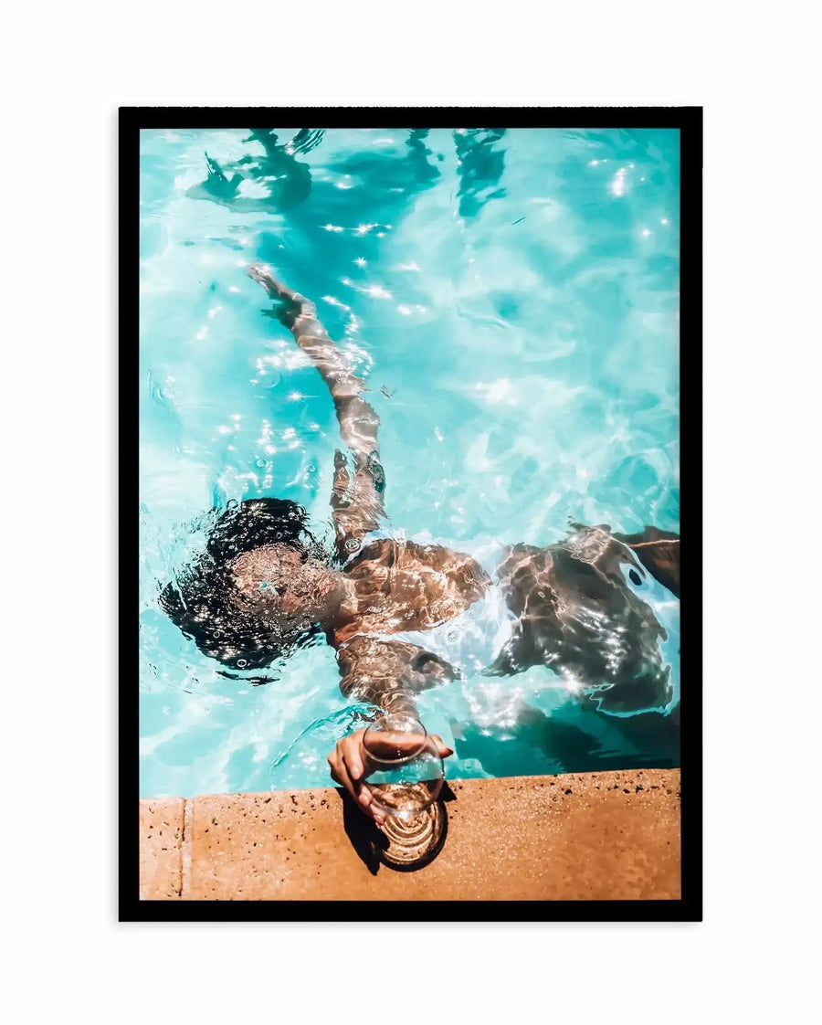 Framed photography art print featuring a woman floating in a bright turquoise swimming pool, viewed from above. Her head and arms are visible at the bottom left, with her body stretching diagonally across the frame. She holds a glass with a drink in her right hand, resting it on the light brown pool edge. Sunlight creates sparkling starburst reflections on the water's surface, and her dark hair is wet and curly. The print is displayed in a black frame with a white border.
