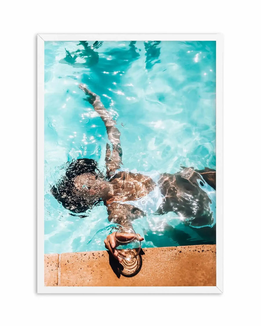 Overhead photograph of a person floating face-up in crystal-clear turquoise water, their dark hair and bronzed skin visible against the luminous blue-green tones. White sunlight sparkles ripple across the water surface. At the bottom of the frame, warm sandy-tan poolside edge with the person's shadow cast across it. The image is framed in white with a clean white border, creating a bright, summery poolside scene with high contrast between the vivid water, golden skin tones, and pale sandy surround.