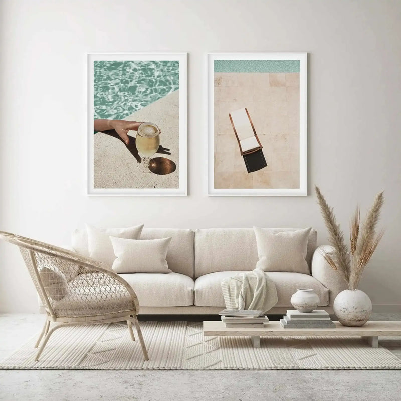 Lifestyle shot of a minimalist living room featuring two framed art prints above a cream sofa. The left print is a photograph of a hand holding a glass of white wine by a swimming pool, with turquoise water and a concrete edge. The right print is an aerial photograph of a poolside scene with a white sunbed on light beige tiles and a strip of turquoise water. The room includes a woven rattan chair, a light wood coffee table with books and ceramic vases, and dried pampas grass.