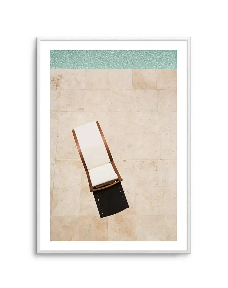 Framed aerial photograph of a poolside scene with a sunbed on beige tiled patio, with white frame and white border. The composition features a light wood and white fabric sunbed with a black towel draped over the foot, positioned on light beige square tiles. A strip of turquoise blue water with ripples is visible at the top of the frame.