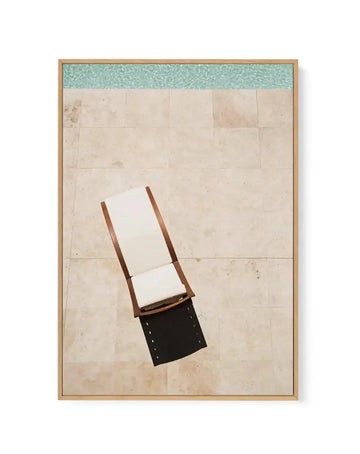 Framed canvas art print featuring an overhead view of a wooden lounge chair with white cushioning and dark seat positioned on a beige tile floor beside turquoise water, rendered in a minimalist illustration style with soft, muted tones and clean geometric shapes, displayed in a natural wood frame with white border.