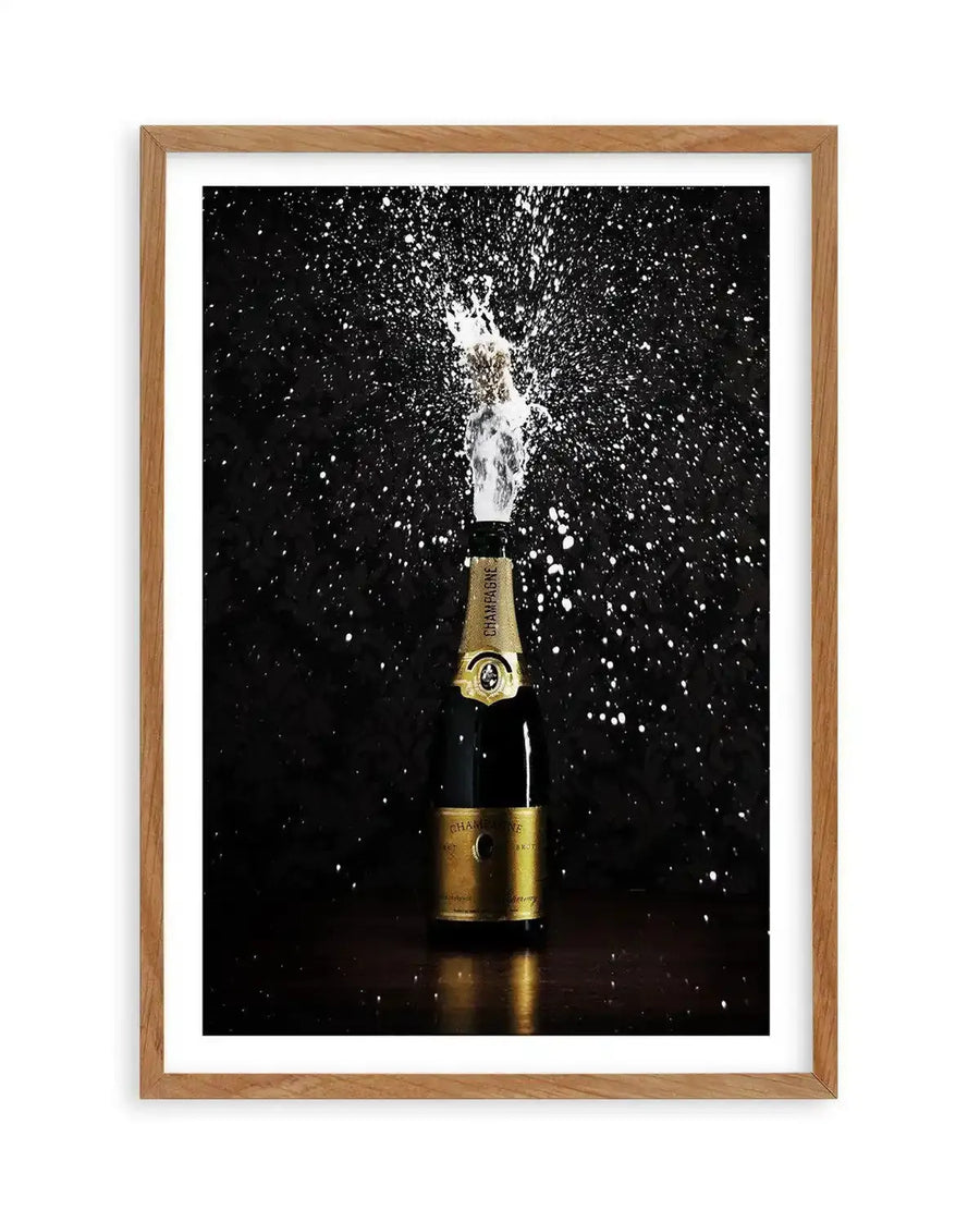"Framed photograph of a champagne bottle popping, with white foam and bubbles spraying outwards against a dark black background, displayed in a natural oak frame with white border. The bottle is gold and black with a gold label, and the spray creates a dynamic, celebratory composition."