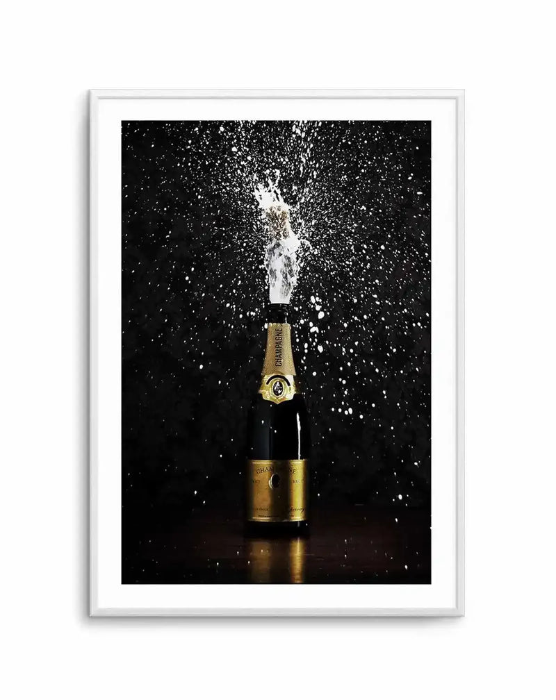 Framed photography art print of a champagne bottle popping, with a stream of white foam and bubbles erupting from the top, set against a dark, almost black background with white splashes. The champagne bottle is gold and dark green, with a gold label, and sits on a dark wooden surface. The print is displayed in a white frame with a white border.