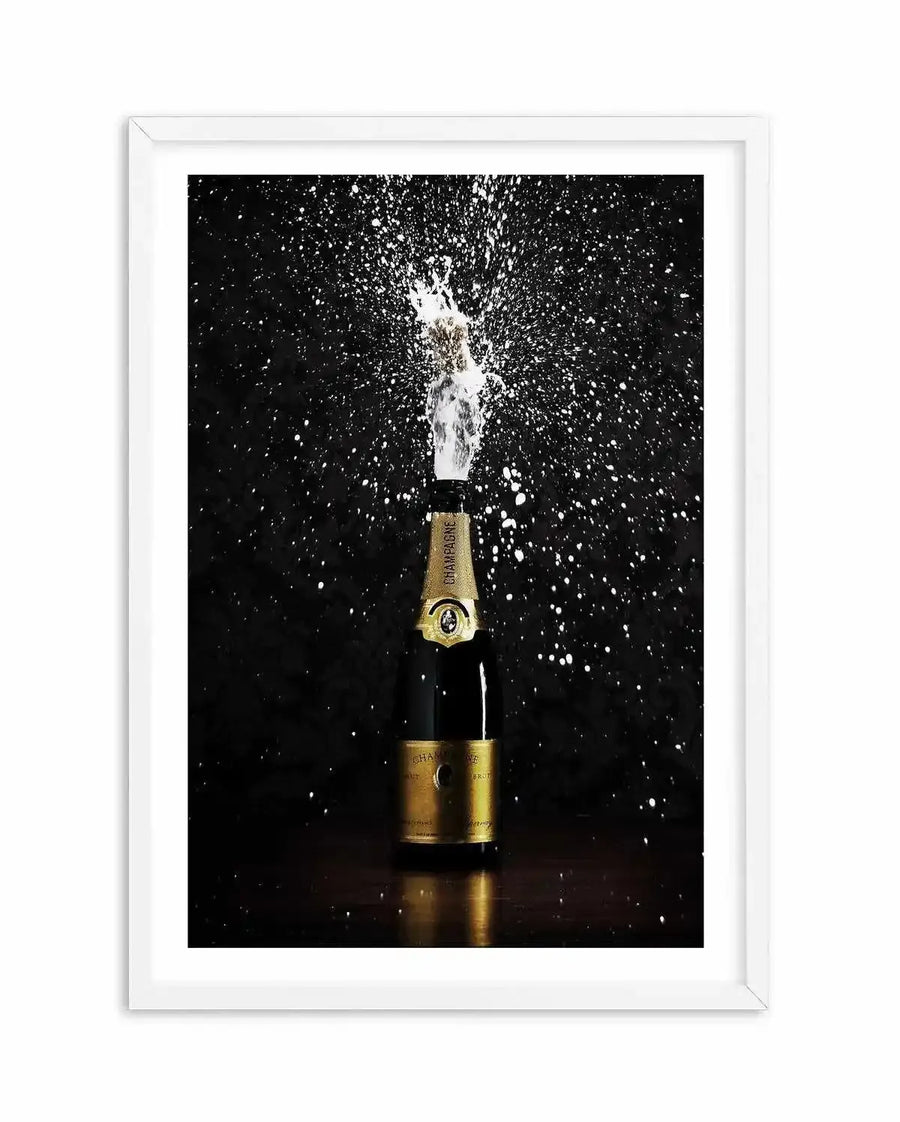 Framed photograph of a champagne bottle popping, with a stream of white foam and sparkling droplets against a dark, almost black background. The bottle is gold and black, reflecting light on a dark wooden surface. The white frame has a white border.