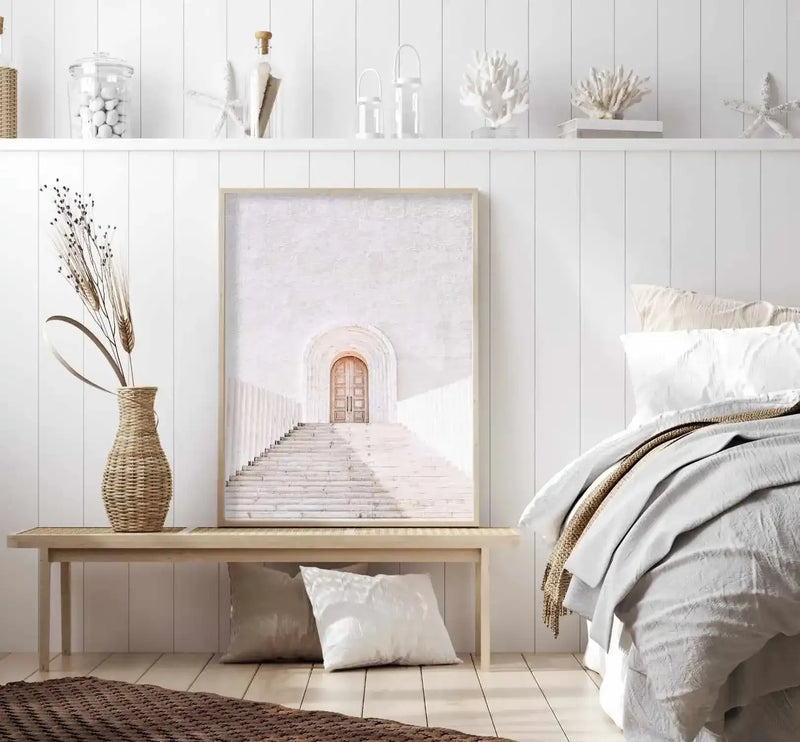 Large framed art print displayed on a light wood bench in a bright, minimalist bedroom with white vertical paneling. The print features a sunlit, white stone staircase leading up to a light wood arched door, creating a serene, architectural composition. The room includes a woven vase with dried grasses, two pillows on the floor, and a bed with white and light grey bedding and a textured brown throw.