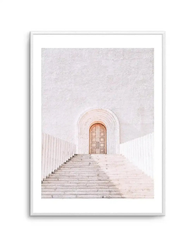 Framed photography art print featuring a minimalist architectural scene in soft white and pale cream tones, with a light wood arched door at the top of a wide stone staircase. The composition is bathed in soft, bright light, creating subtle shadows and a serene mood, displayed in a white frame with a white border.