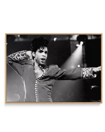 Black and white framed canvas art print featuring a performer in an ornate dark jacket with decorative white beading down the front and patterned white designs on the cuffs, arms raised in an expressive pose against a dark background with stage lighting. The photograph is mounted in a natural wood-tone frame with clean lines.